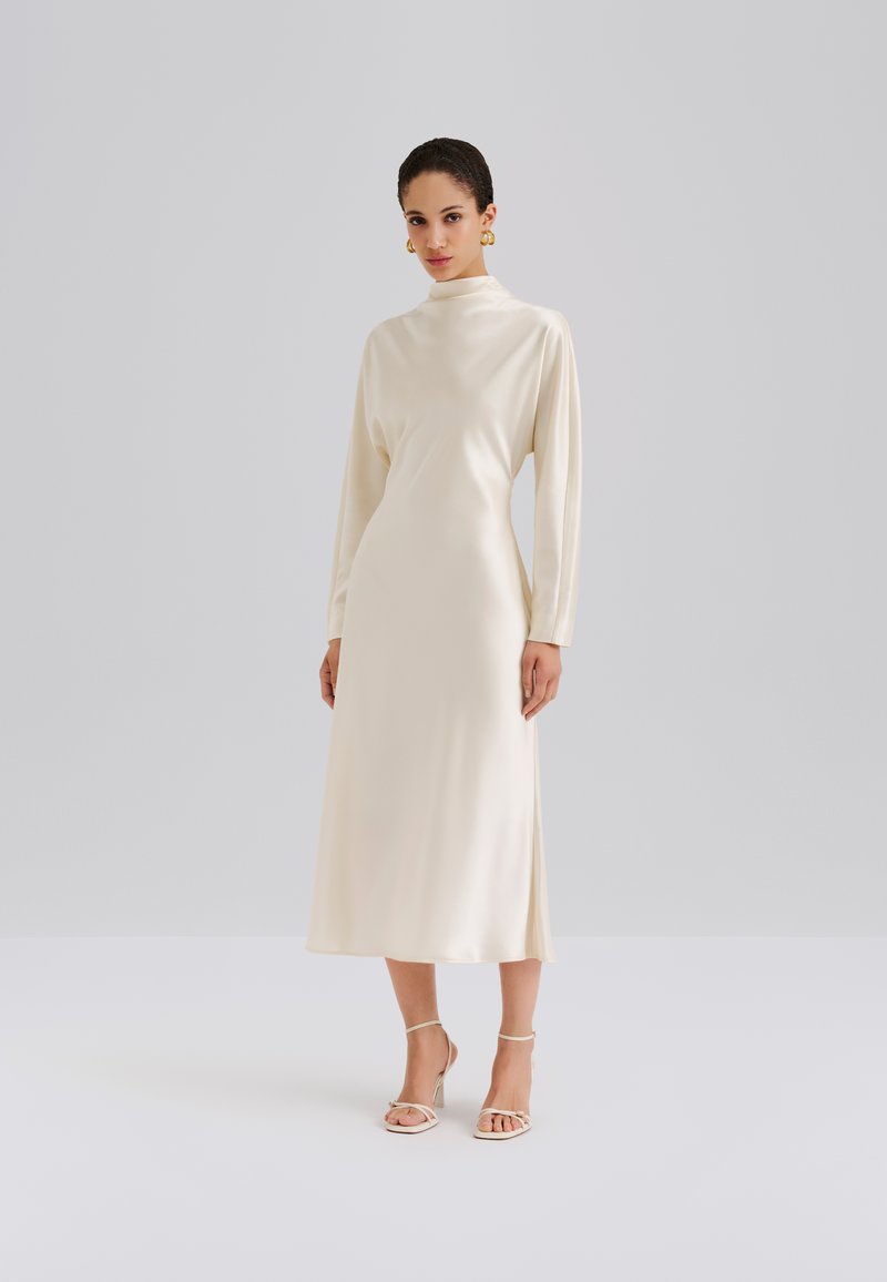 Vestido de satén color crema con mangas largas, cuello alto y silueta en línea A. Textura suave y detalle de abertura lateral. Combinado con tacones de tiras.