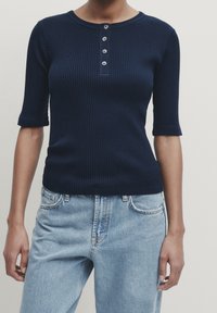Marineblå ribbet kortærmet top med rund hals og fem dekorative knapper ned foran. Kombination med lyseblå jeans.