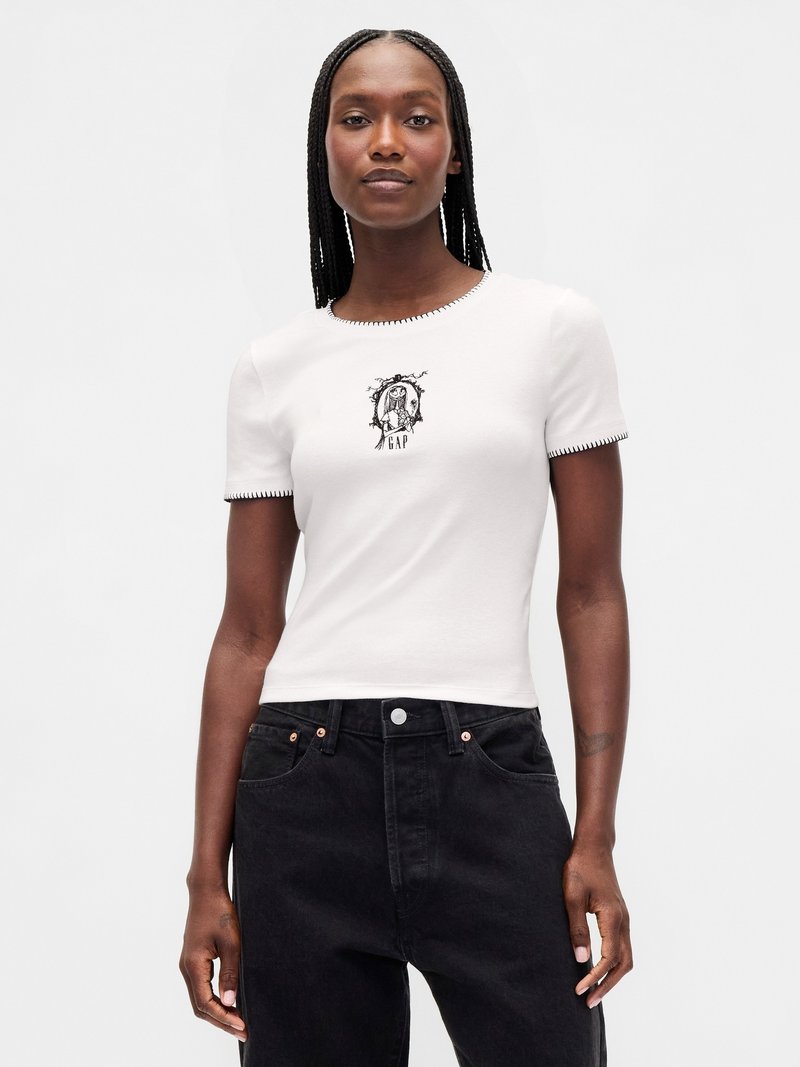 Mulher com cabelo trançado, vestindo uma blusa branca de mangas curtas com costuras pretas e um pequeno gráfico preto, combinada com jeans pretos de cintura alta.