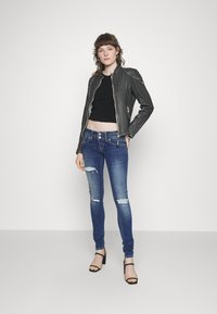 Chaqueta de cuero negra con cierre de cremallera, combinada con vaqueros ajustados azul oscuro con detalles desgastados y tacones negros con tiras.