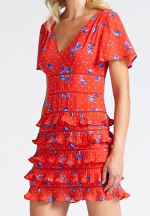 Kobieta ubrana w czerwoną sukienkę w kwiaty z niebieskimi kwiatami, białymi kropkami, krótkimi rękawami, dekoltem w serek oraz wielowarstwową falbanianą spódnicą.