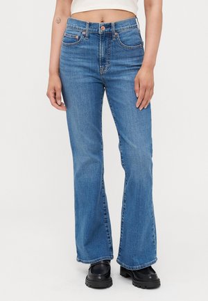 Žena v modrých vysokým pasem rozšířených džínách, černých masivních botách a bílém zkráceném topu stojící na jednobarevném pozadí.