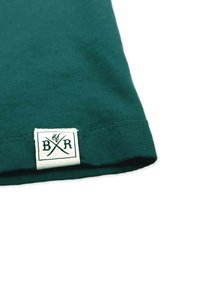 Tummanvihreä puuvillapaita, jossa on valkoinen kangastarra, jossa lukee "BXR" ja ristikkäisiä viivoja. Liukas pinta, yksinkertainen muotoilu, ei kuvioita.