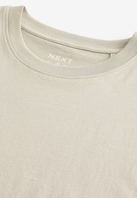 Tricou din bumbac bej deschis, cu guler rotund. Are o textură moale și un model subtil, etichetă de marcă cosută în interiorul gulerului.