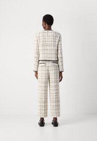 Costume à carreaux beige avec une veste courte et un pantalon évasé. Présente des garnitures noires contrastantes et un tissu texturé. Poches arrières plaquées sur le pantalon.