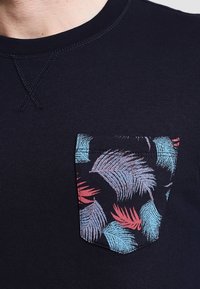 Navyblaues Shirt mit Rundhalsausschnitt und einer bunten Brusttasche im Muster von Palmenblättern in Blau-, Rot- und Violetttönen.