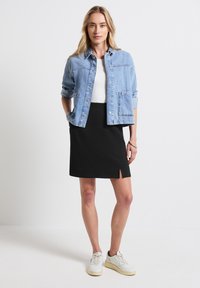 Veste en denim bleu clair avec fermeture à boutons, deux poches avant ; associée à une jupe noire ajustée avec une fente sur le devant. Baskets blanches.