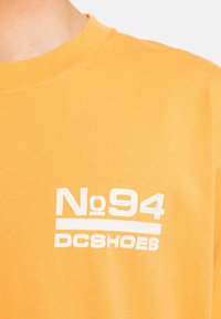 Short-sleeve, round-neck t-shirt in bright yellow cotton. Features a white graphic text "No 94 DC Shoes" on the front. Smooth texture.