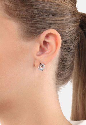 Orecchio di donna con capelli castano chiaro raccolti, che indossa un piccolo orecchino a forma di cuore in argento con pietre trasparenti incastonate.