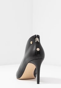 Chaussure à talon haut en cuir noir avec un bout pointu, une fermeture éclair à l'arrière et des accents de quincaillerie finition or. Texture lisse et design élégant.