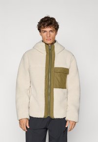 Tormekas jakk off-white värvuses, mille eesmine paneel ja taskud on oliivrohelised. Omab lukuga sulgemist ja kapuutsi. Tekstuurne ja mugav välimus.