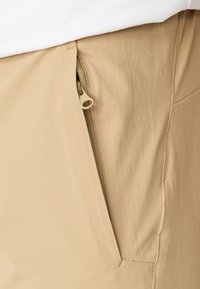 Pantalons beiges présentant une poche latérale avec fermeture éclair. Le tissu semble lisse, avec une texture mate et des détails de couture subtils.