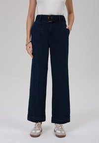 Högmidjade, vida denimbyxor i mörkblått med ett framträdande bälte, två fickor, synlig sömnad och uppvikt fåll.