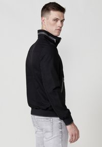 Chaqueta bomber negra con un alto cuello, frente con cremallera y puños acanalados. Presenta un diseño aerodinámico y una textura elegante.