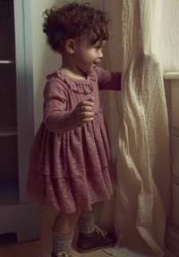Różowa sukienka warstwowa z teksturowanej tkaniny, z falbanistym kołnierzem i długimi rękawami. Dziecko nosi wełniane skarpetki i brązowe buty, stojąc przodem do zasłony.