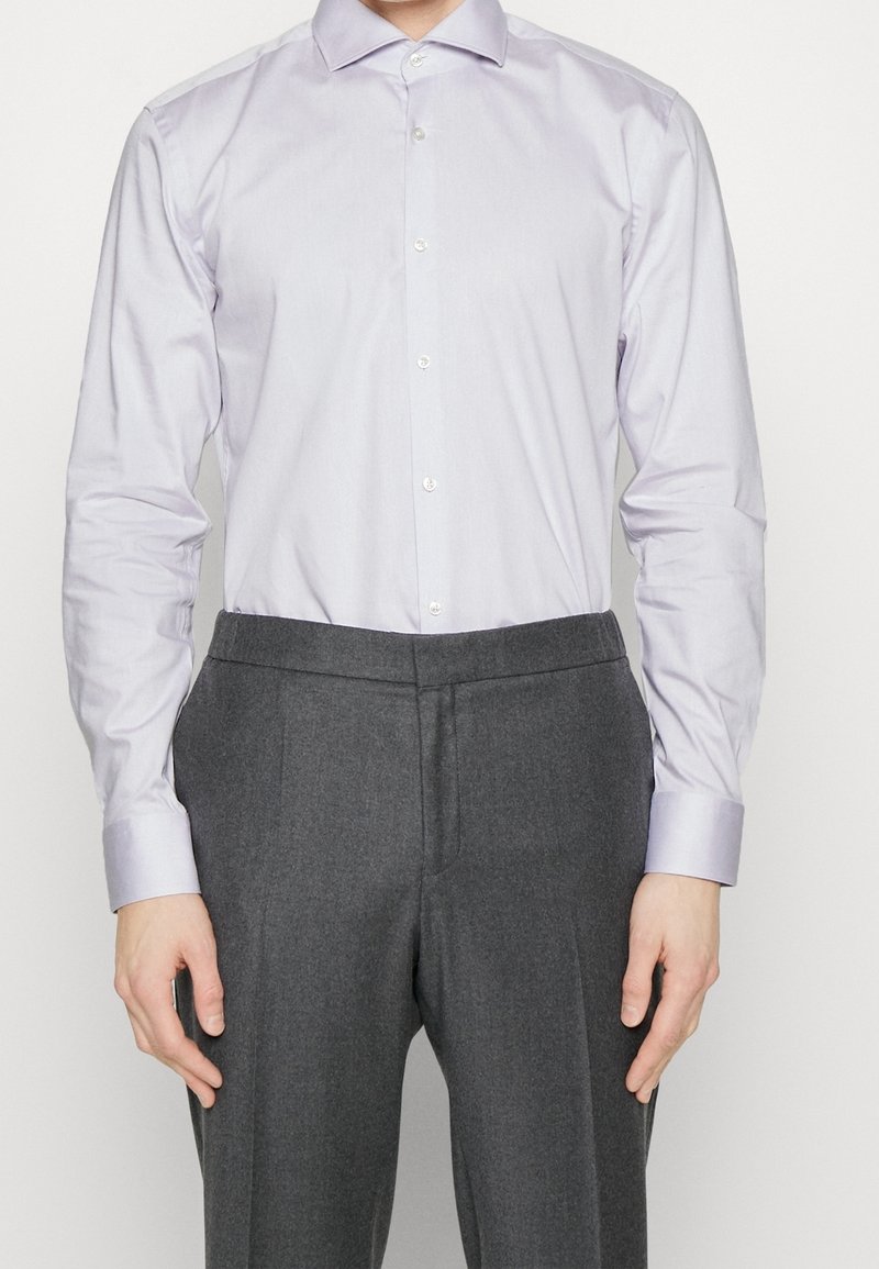 Chemise à manches longues de couleur violet clair avec un col pointu et une fermeture par boutons, associée à un pantalon tailleur gris foncé. Texture de tissu lisse.
