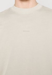 T-shirt a maniche lunghe beige in tessuto morbido, caratterizzato da un collo a girocollo a coste e un logo in rilievo di una tonalità leggermente più chiara sul fronte.