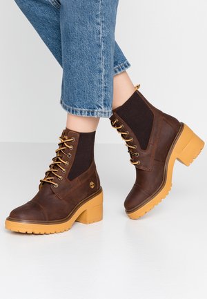 Bottes montantes en cuir marron à lacets avec talons épais jaunes, portées avec un jean bleu court sur un sol blanc.