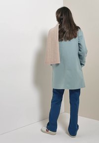 Persona con lunghi capelli castani sta in piedi di fronte all'angolo indossando un cappotto azzurro chiaro, pantaloni blu scuro, sneakers bianche e una sciarpa beige sulla spalla.