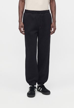 Mand iført sorte sweatpants med snøre og sorte sneakers med hvide striber, står mod en ensfarvet hvid baggrund.