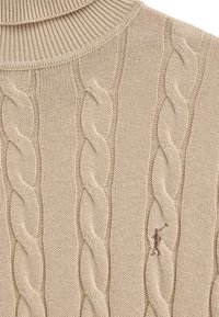 Beige stickad tröja med ribbad polokrage och texturerat kabelmönster. Har en liten broderad figur i en kontrastrik brun tråd.
