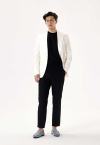White blazer over a black shirt, paired with black trousers and light blue loafers. Smooth fabrics and tailored design highlight the ensemble.