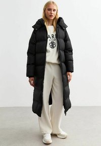 Abrigo negro tipo puff con cierre frontal y capucha, que presenta secciones acolchadas. Llevado sobre un suéter crema y pantalones a juego, de pie sobre un suelo gris.