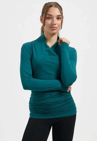 Ung kvinna med håret uppsatt som bär en turkos långärmad topp med rynkor och svarta byxor, poserande med en hand nära halsen.