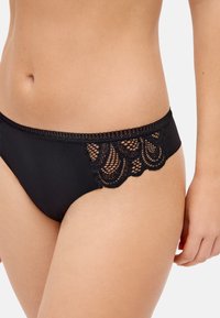 Ropa interior estilo bikini negra con detalles de encaje y malla en las caderas, usada por una persona sobre un fondo liso.