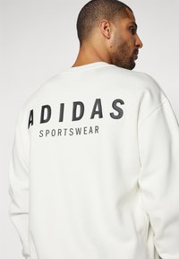 Balta džemperis ar apaļu kakla izgriezumu, gluda auduma un liela melna uzraksta "ADIDAS SPORTSWEAR" aizmugurē.