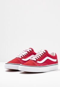 Paire de baskets basses rouges avec lacets blancs, bande latérale blanche et semelles blanches épaisses sur un fond clair uni.