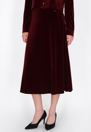 Modelo con una falda midi de terciopelo color burdeos, una chaqueta a juego y zapatos negros de tacón alto y punta fina, sobre un fondo blanco liso.