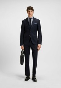 Abito blu con giacca aderente e pantaloni dritti. Indossato con una camicia a quadri chiara, cravatta e un fazzoletto blu. Scarpe e borsa in pelle nera.
