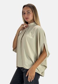 Camicia beige oversize con collo alto, maniche corte e tessuto texturizzato. Abbinata a jeans neri in un'angolazione laterale.