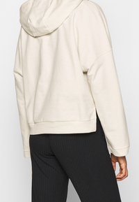 Sudadera corta beige con un acabado suave y texturizado, con capucha y hombros caídos, combinada con pantalones negros de canalé.