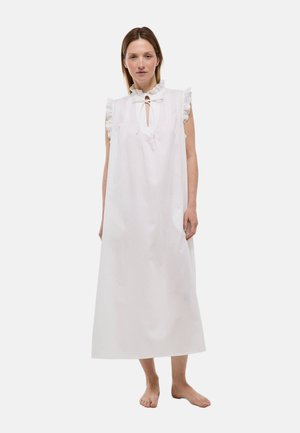 Femme debout pieds nus portant une longue robe blanche sans manches avec des épaules à volants et un col en trou de serrure noué.