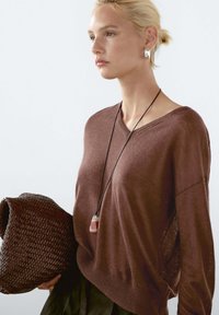 Brązowy sweter dzianinowy z dekoltem w serek, luźny krój i boczne rozcięcia. W połączeniu z plecioną torbą w ciemnym brązie i naszyjnikiem z zawieszką.
