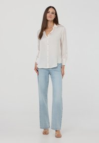 Camicia bianca a maniche lunghe con colletto, abbinata a jeans azzurri a gamba larga. Il modello è in piedi a piedi nudi su uno sfondo neutro.