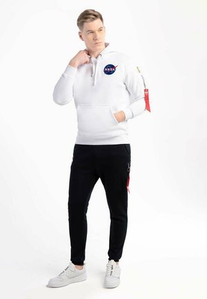 White NASA hoodie with drawstrings, front pocket, and logo; paired with black pants featuring zippered pockets. Worn with white sneakers.