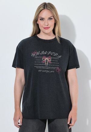 Blondynka w luźnej czarnej koszulce z ozdobnymi kokardkami i białym, kursywnym napisem, w studio na jasnym tle.