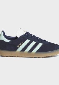 Σκούρο μπλε αθλητικό παπούτσι Adidas Gazelle με λευκές ρίγες, λαστιχένια σόλα και χρυσή επιγραφή λογότυπου στην πλευρά, ορατό από έξω.