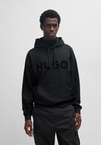 Schwarzer Hoodie mit einem großen, glänzenden "HUGO"-Logo auf der Vorderseite, lockerer Schnitt, verstellbare Kapuze mit Kordelzug und vorderer Kängurutasche.