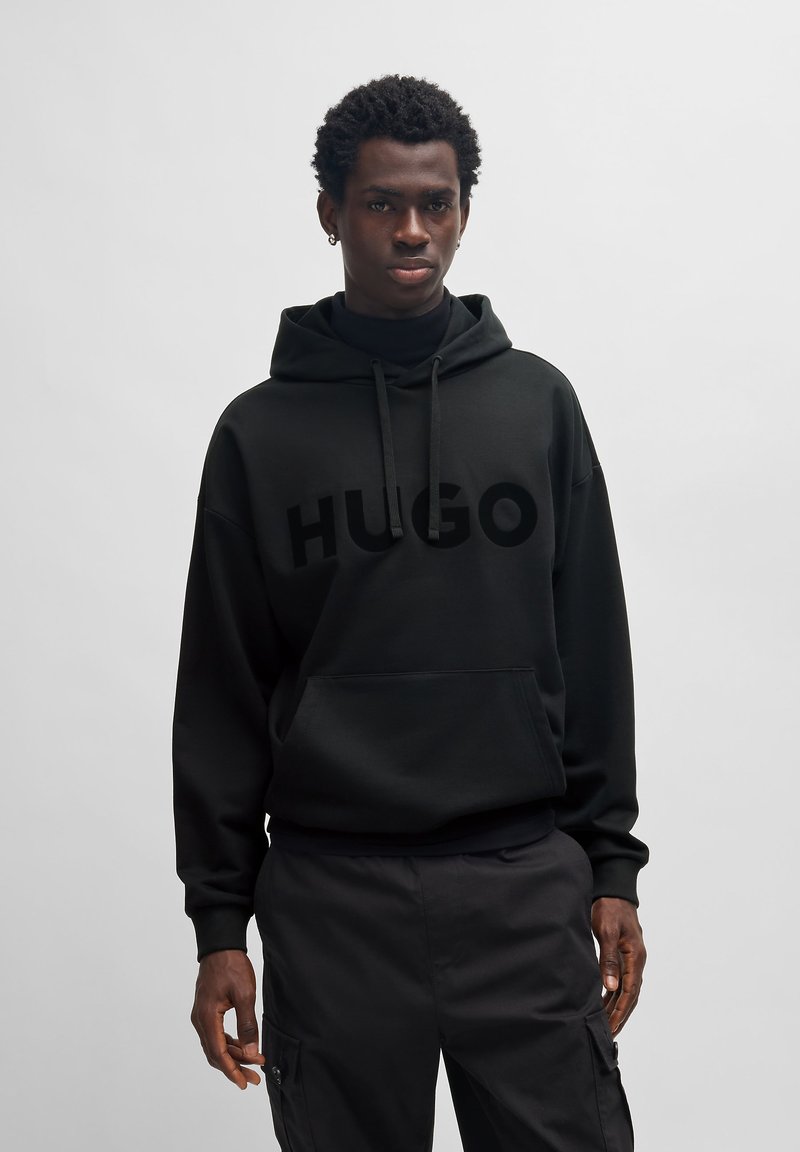 Schwarzer Hoodie mit einem großen, glänzenden "HUGO"-Logo auf der Vorderseite, lockerer Schnitt, verstellbare Kapuze mit Kordelzug und vorderer Kängurutasche.