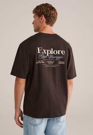 Braunes Baumwoll-T-Shirt mit kurzen Ärmeln, das auf der Rückseite weißen Text mit der Aufschrift "Erkunde deinen Horizont" und weiteren beschreibenden Phrasen zeigt.