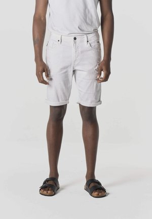 Hombre con shorts de mezclilla blancos remangados, camiseta blanca y sandalias negras, de pie contra un fondo claro y liso.