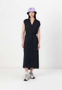 Abito senza maniche nero con bottoni e cintura da annodare in vita, caratterizzato da una silhouette dritta. Completato da un cappello a secchiello viola chiaro e sneakers.