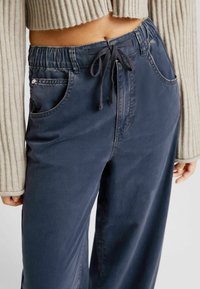 Blå bomullshängselbyxor med elastiskt midjeband och dragsko, med sidofickor och avslappnad passform. Texturerad ribbad ärm synlig.