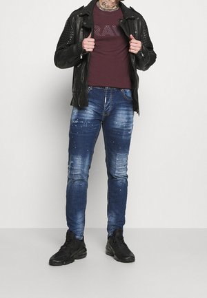 Veste de moto en cuir noir avec accents aux épaules, t-shirt bordeaux, jean bleu déchiré avec éclaboussures de peinture, et baskets noires.