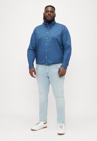 Modrá košeľa s gombíkmi vyrobená z bavlny, v kombinácii so svetlomodrými džínsami. Má golier a voľný strih. Biely tenisky s hnedými akcentmi.