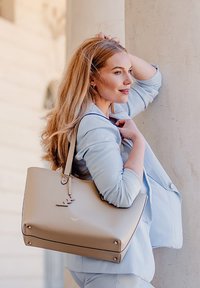 Béžová taška přes rameno s elegantním designem, hladkou texturou a výraznými kovovými akcenty na dně. Nosit ji na rameni na světlém pozadí.